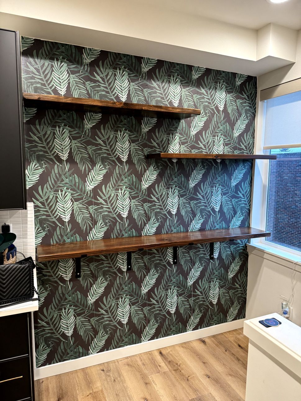 Three wooden shelves on a wall covered with tropical patterned wallpaper. Black brackets support shelves.