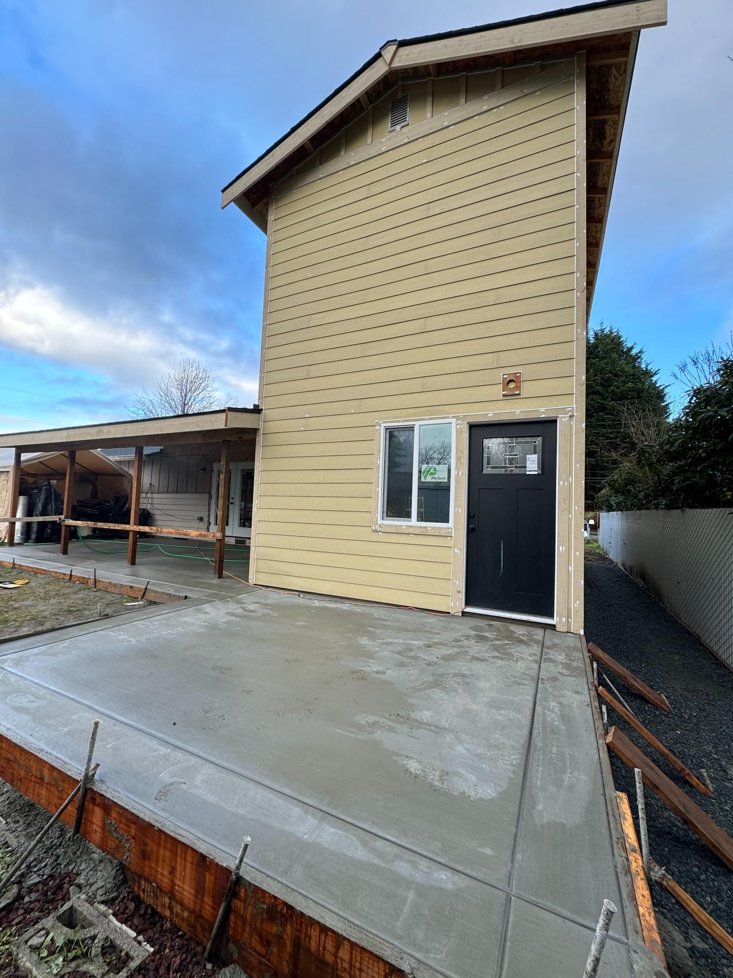 Exterior view of a newly constructed house with a concrete patio. Yellow siding, black door, cloudy sky.