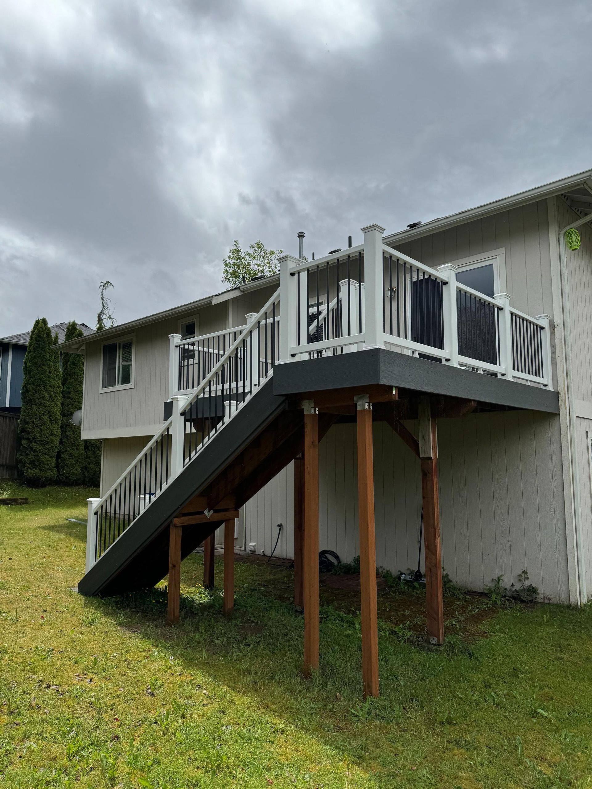 Elevated deck with white railing, gray decking, and brown support posts attached to a light-colored house under a cloudy sky.