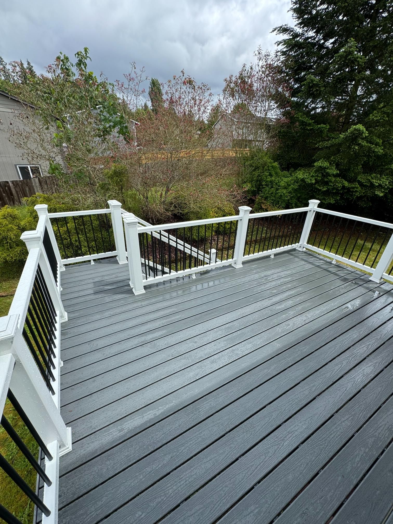 Gray composite deck with white railings and black balusters, overlooking a backyard with trees under a cloudy sky.