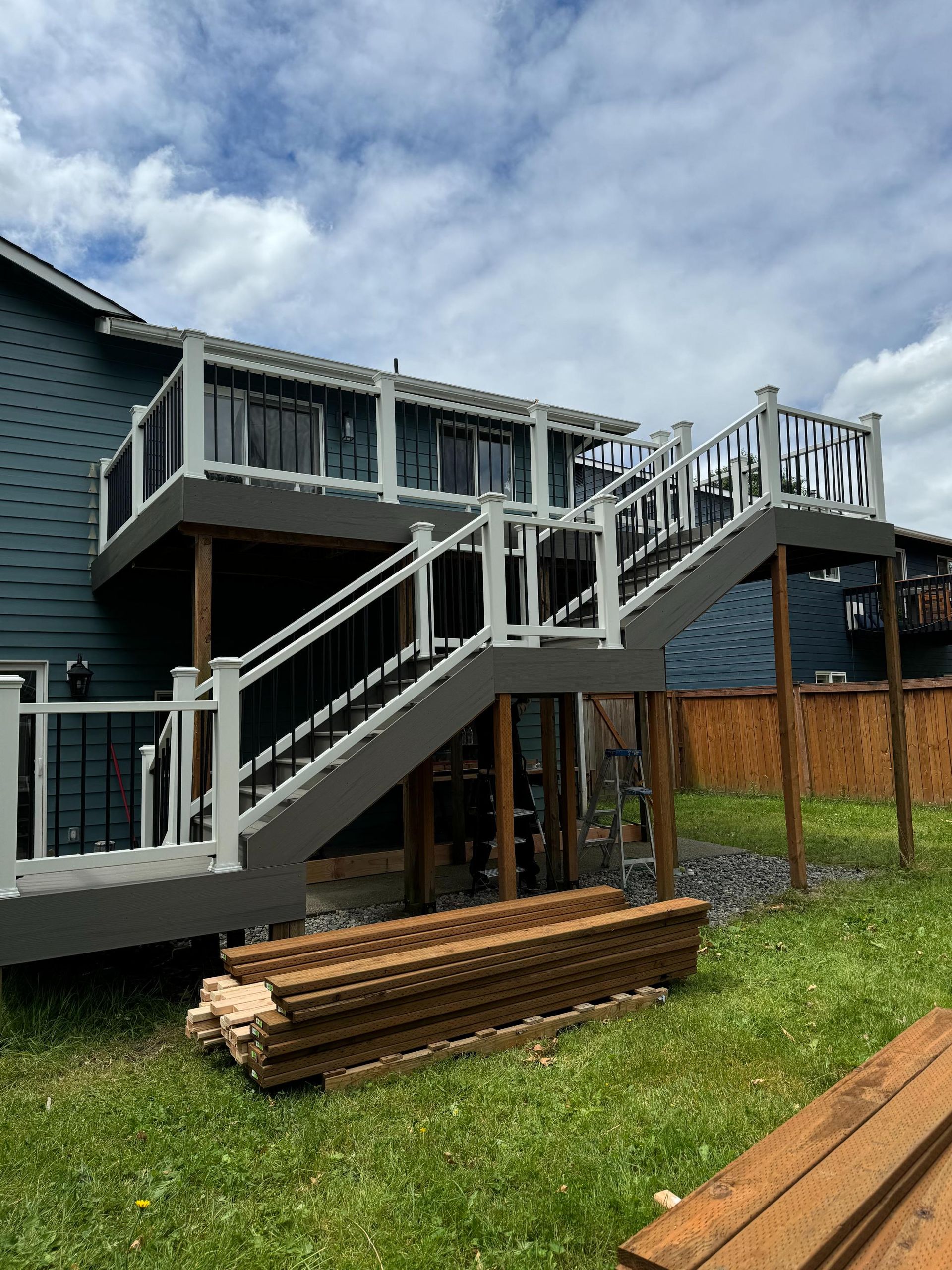 Two-level deck with stairs; gray and white railings, black mesh, brown support beams, stacked lumber on grass.