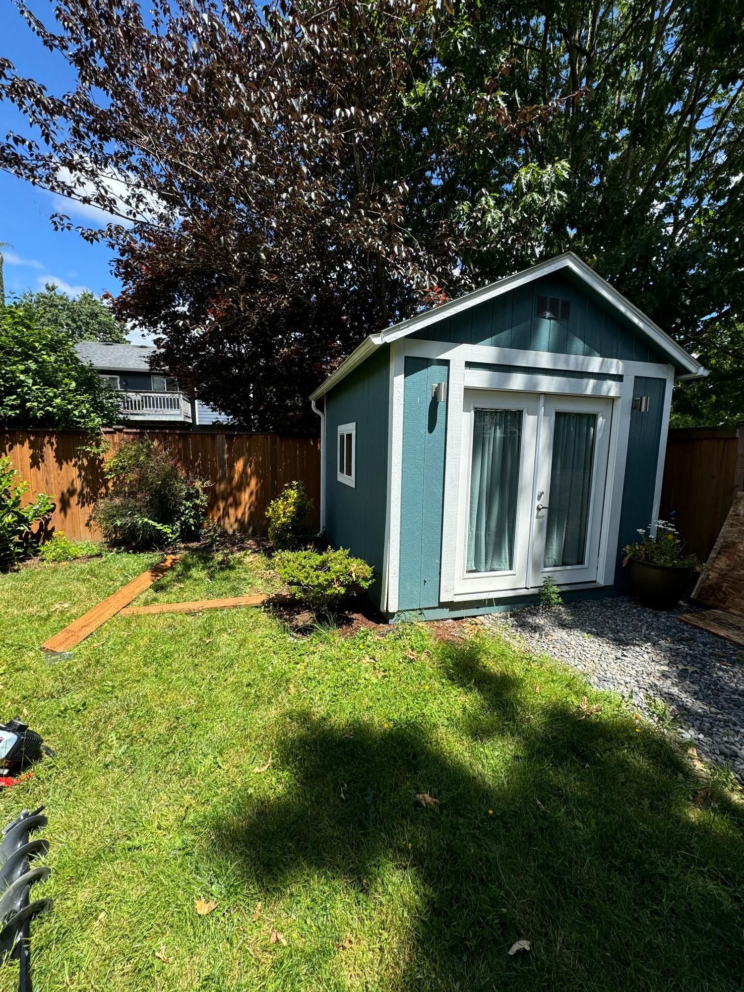 Teal shed with white trim, double doors, and small window in a backyard with green grass, gravel, and a brown fence.