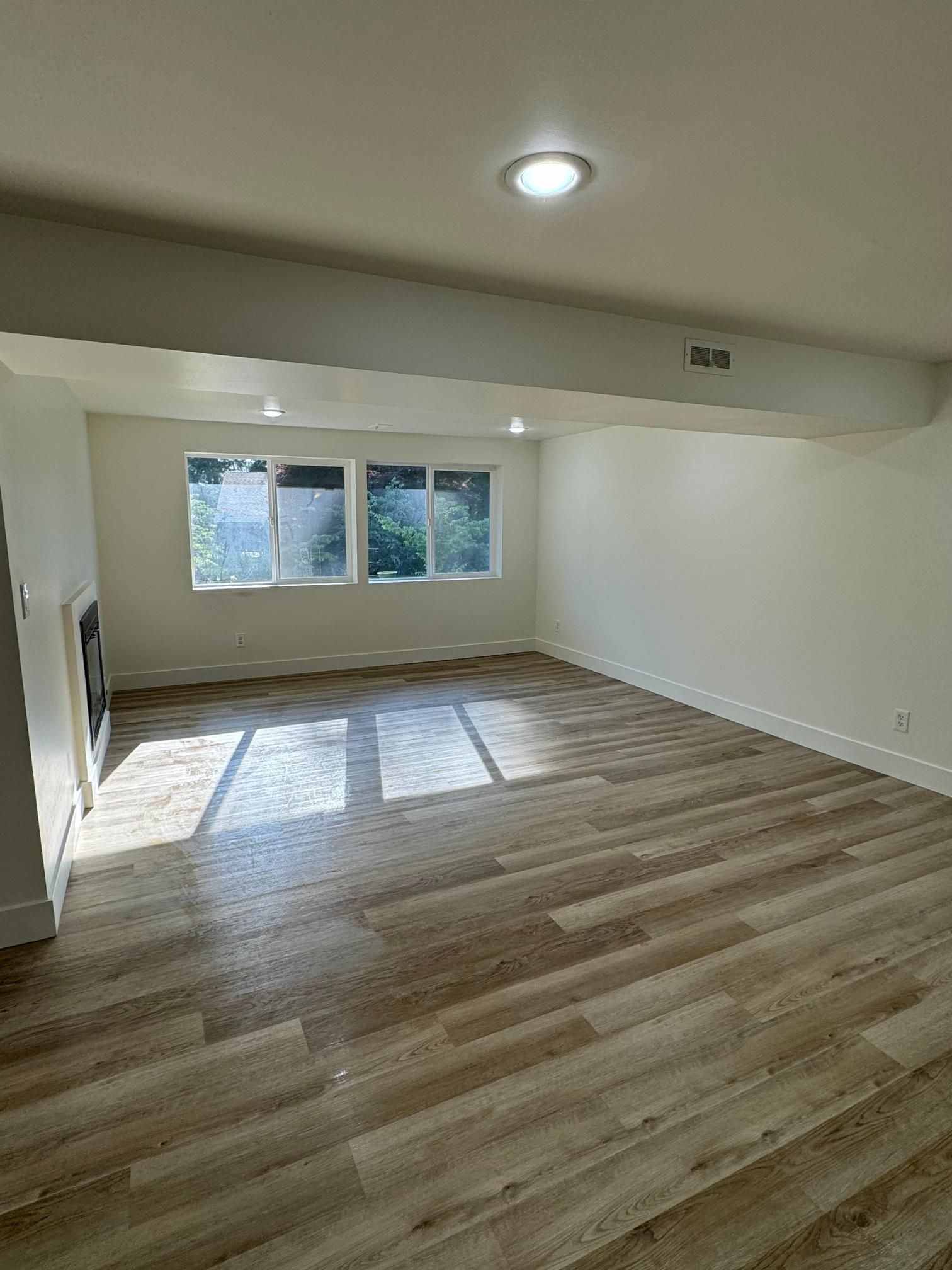 Empty, bright room with wood-look flooring, white walls, three windows with sunlight, and a recessed ceiling light.