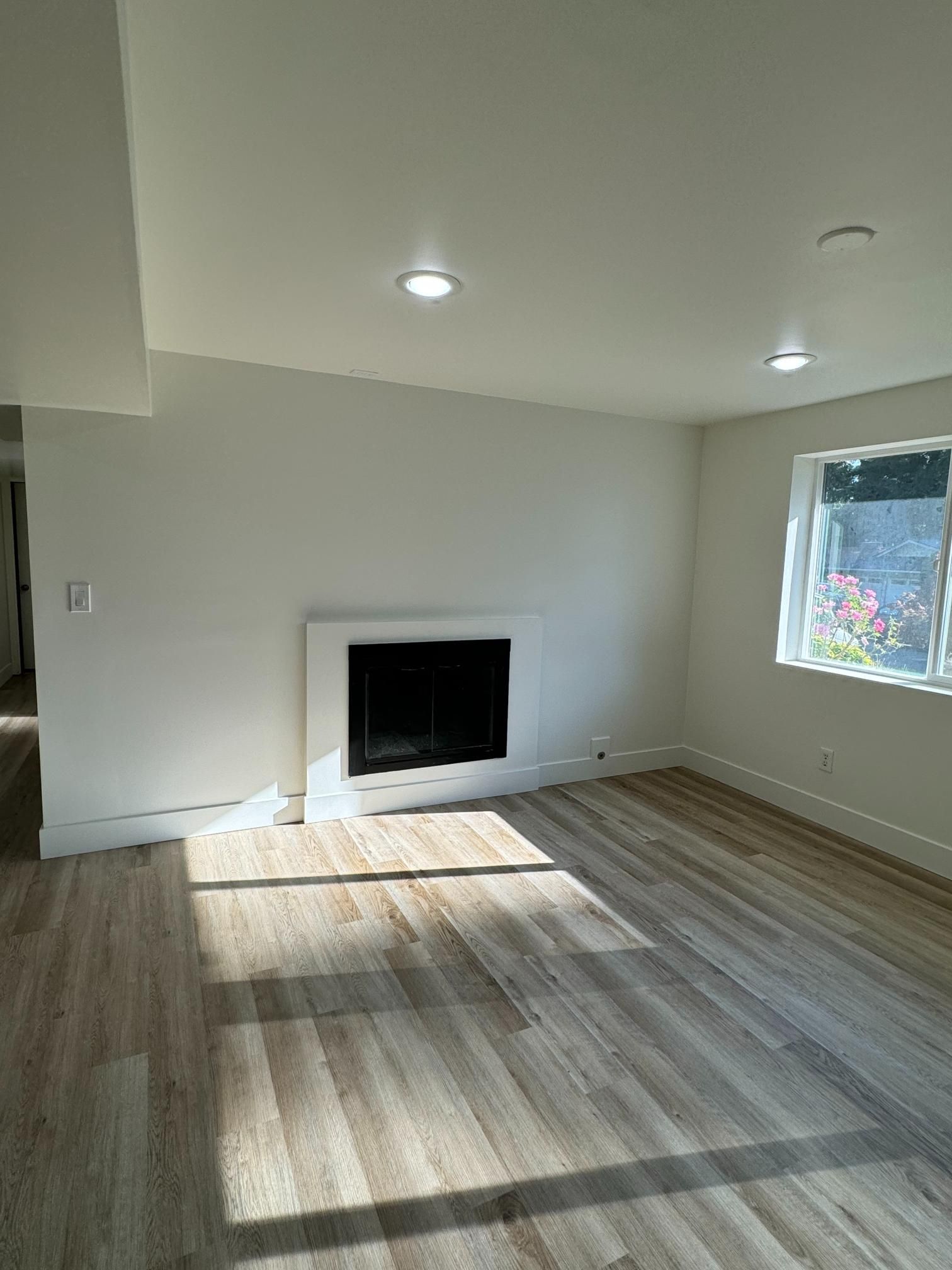 Empty living room with fireplace, white walls, and wood-look flooring. Sunlight streams through the window.