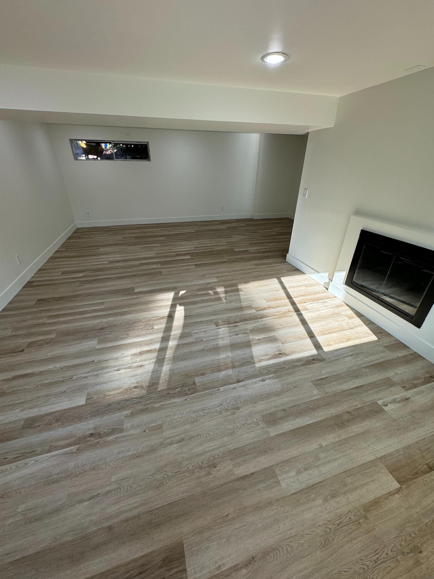 Empty room with wood-look flooring, white walls, and a fireplace. A small window lets in light.