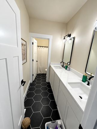 Narrow bathroom with black hex tile floor, double white vanity, and two mirrors against beige walls.