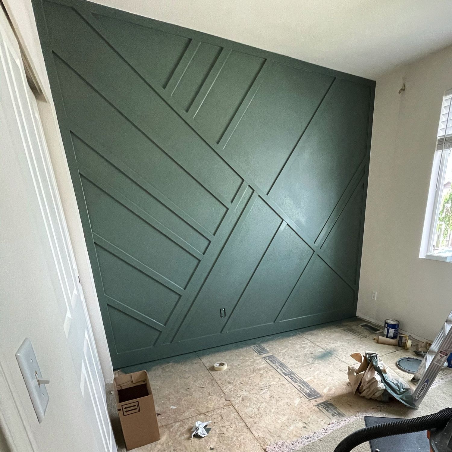 Green geometric accent wall under construction in a room with a window and a door.