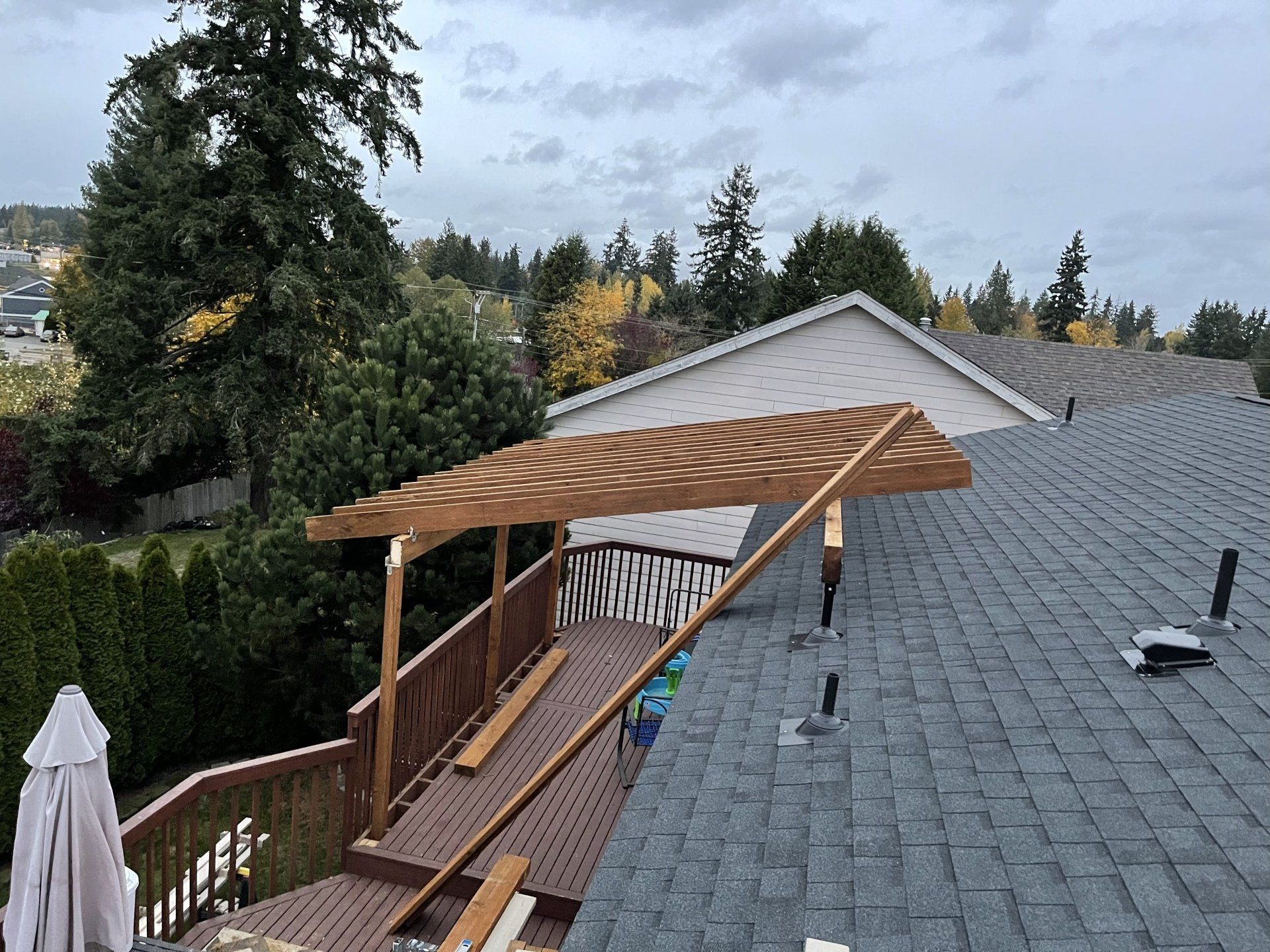 Wooden deck with pergola attached to a house with gray shingles and surrounded by trees under a cloudy sky.