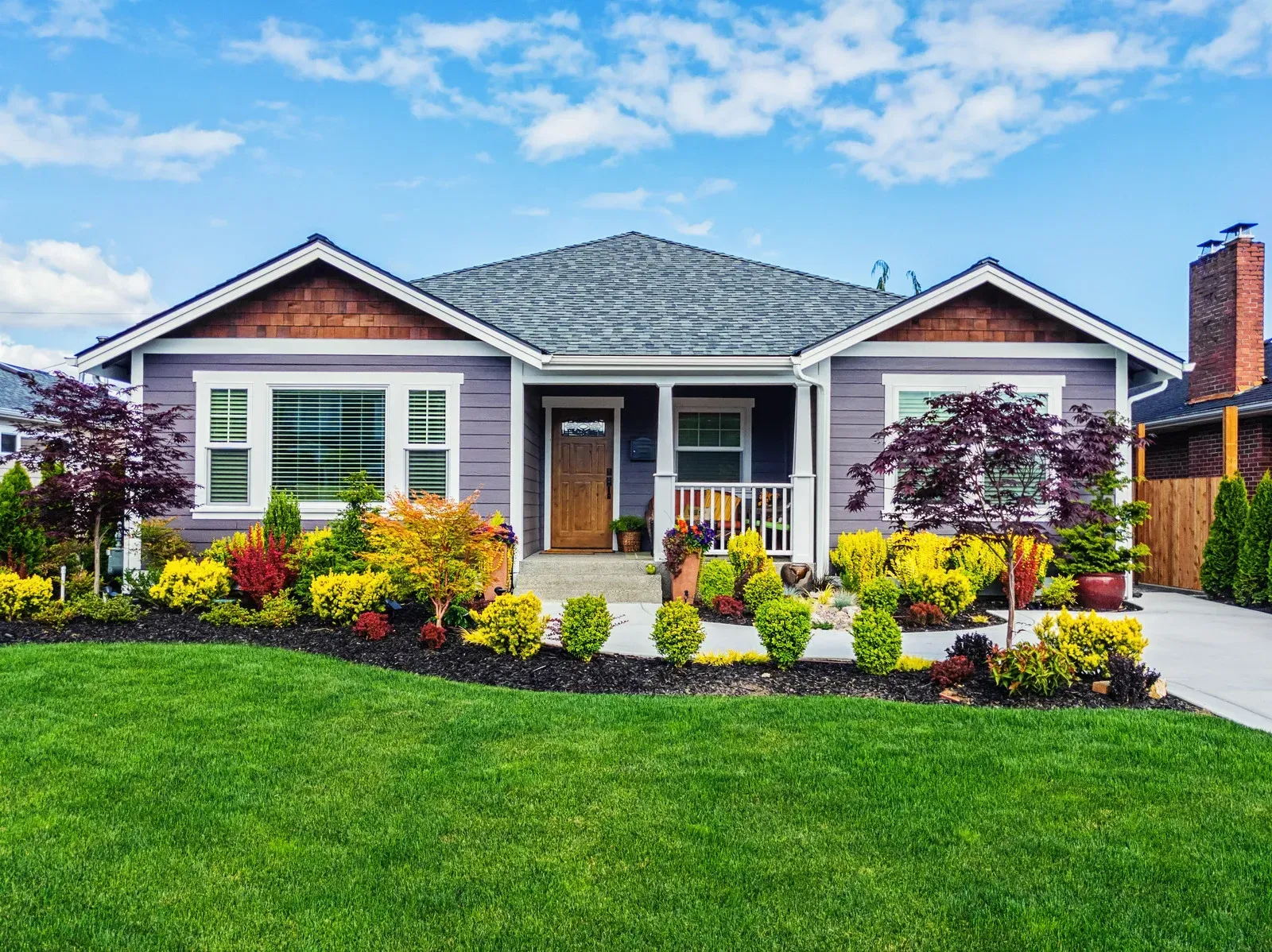 Single-story gray house with white trim, colorful front garden, and lush green lawn under a blue sky