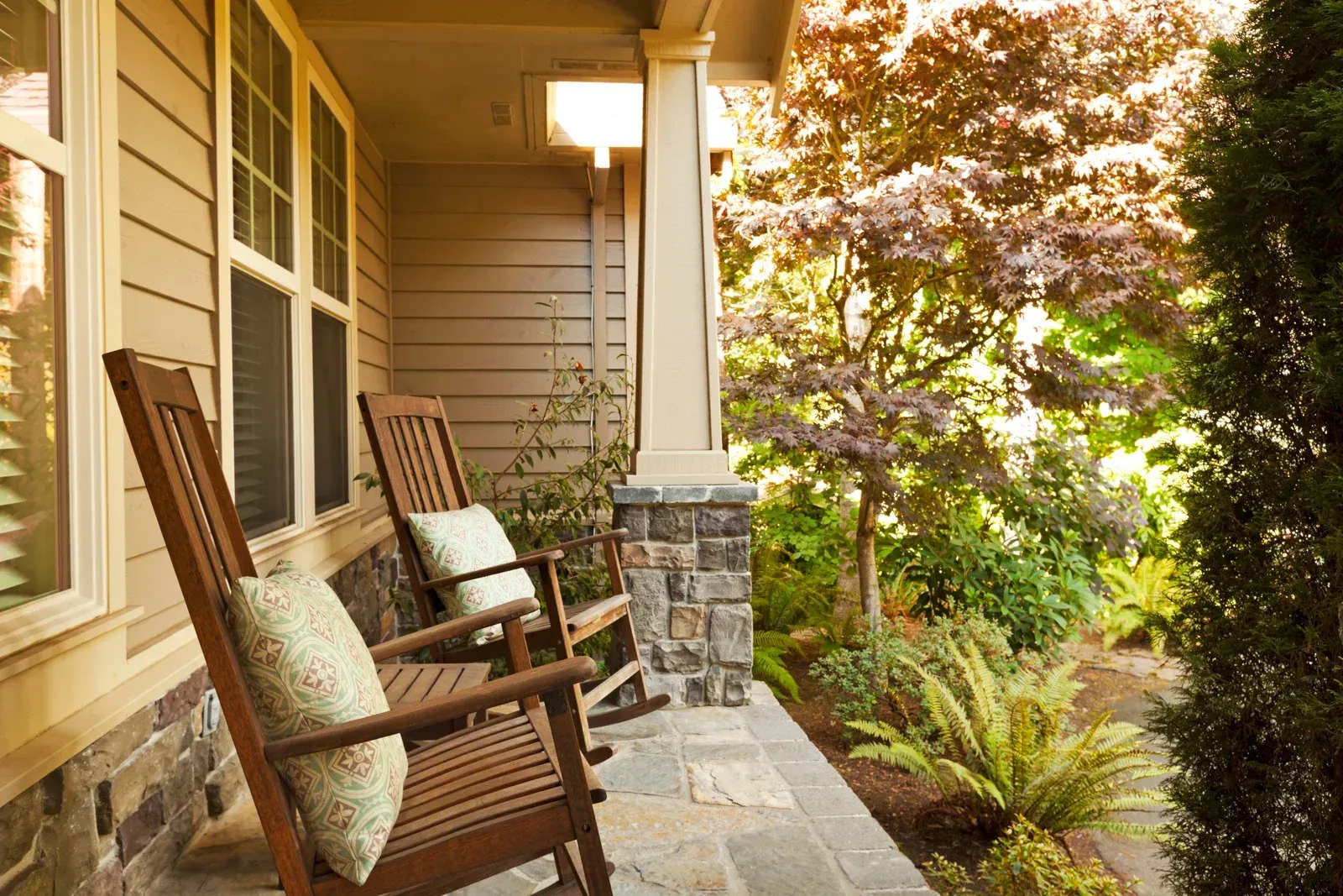 Front porch with wooden chairs and cushions beside a stone column, overlooking a leafy garden.