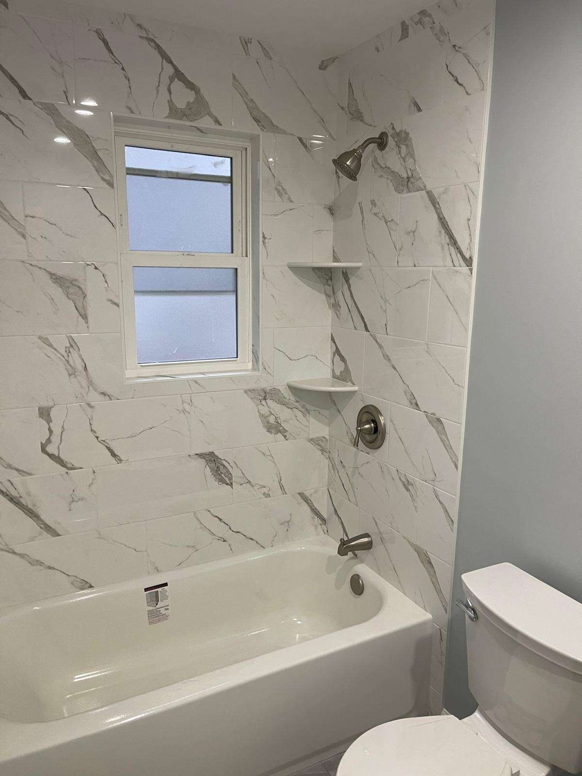 Small bathroom with marble-look shower wall, tub, window, shelves, and toilet.