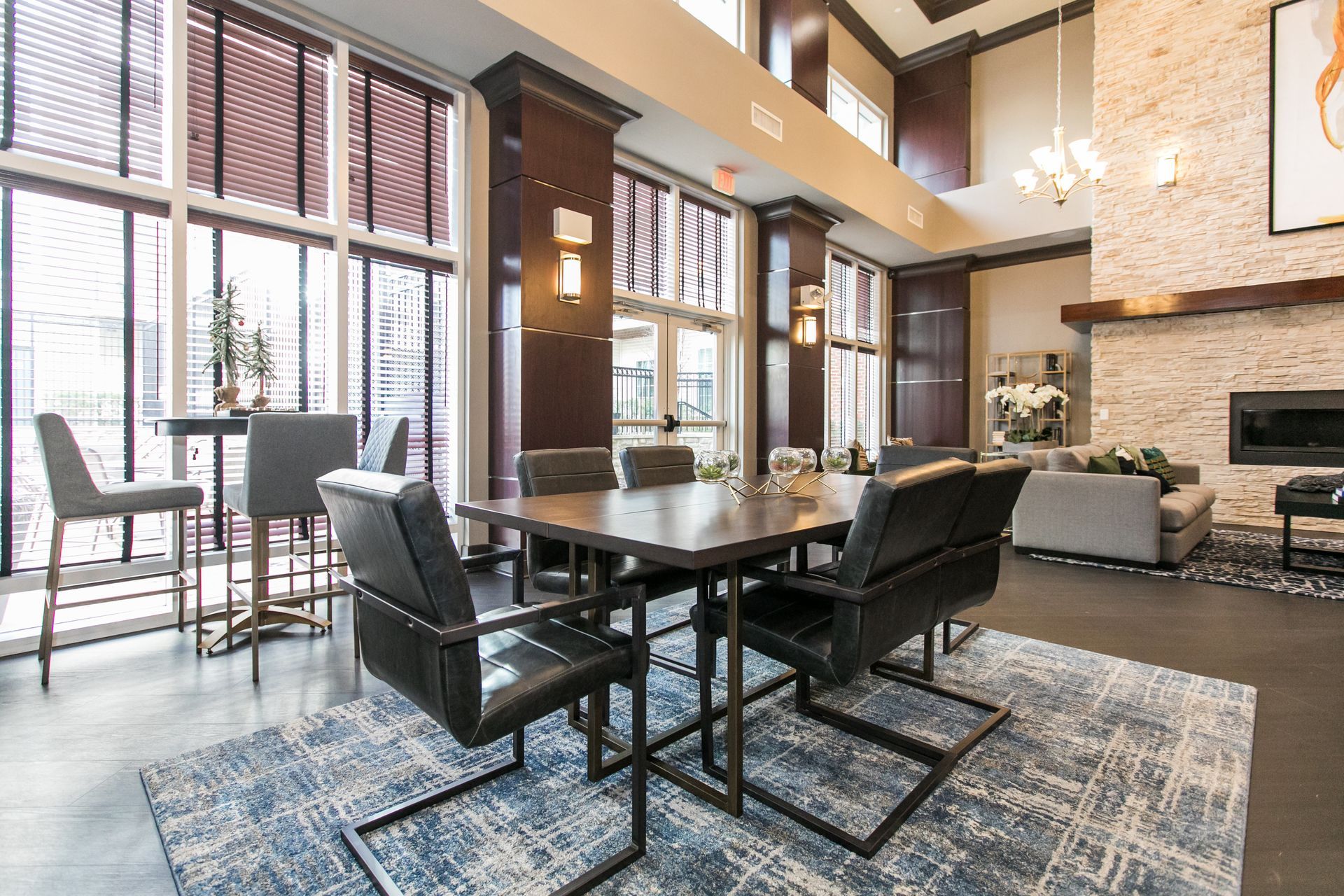 Modern dining room with large windows, black chairs, a long table, and a stone fireplace in warm tones at Verde at Howard Square West Apartments in Elkridge, MD.