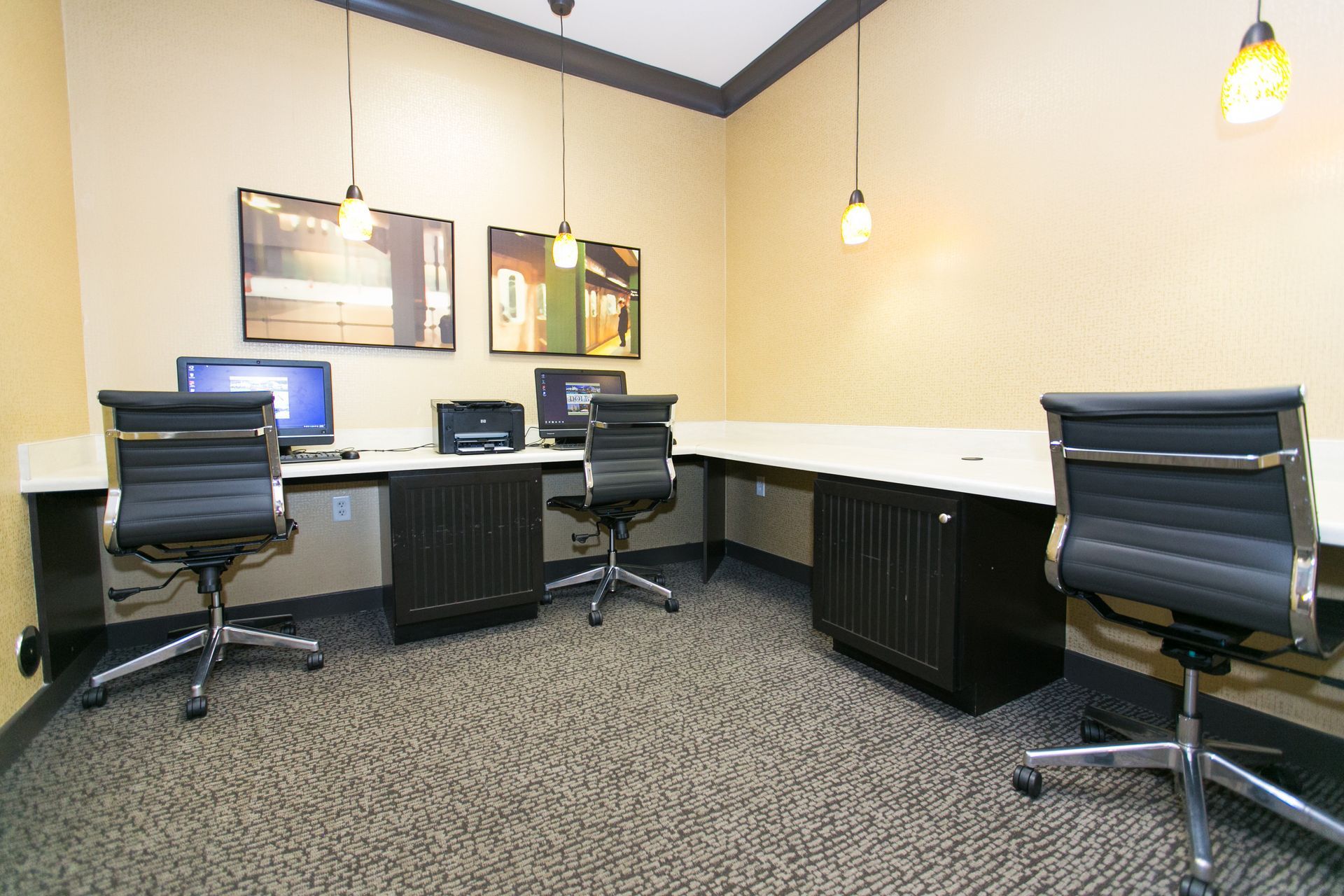 Modern office workspace with desks, computers, black chairs, and pendant lights under beige walls at Verde at Howard Square West Apartments in Elkridge, MD.
