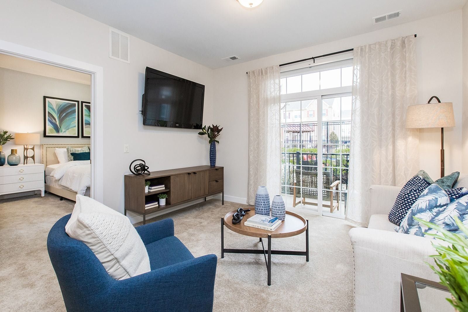 Bright living room with blue chair, wall-mounted TV, glass coffee table, sofa, and French doors to a patio at Verde at Howard Square West Apartments in Elkridge, MD.
