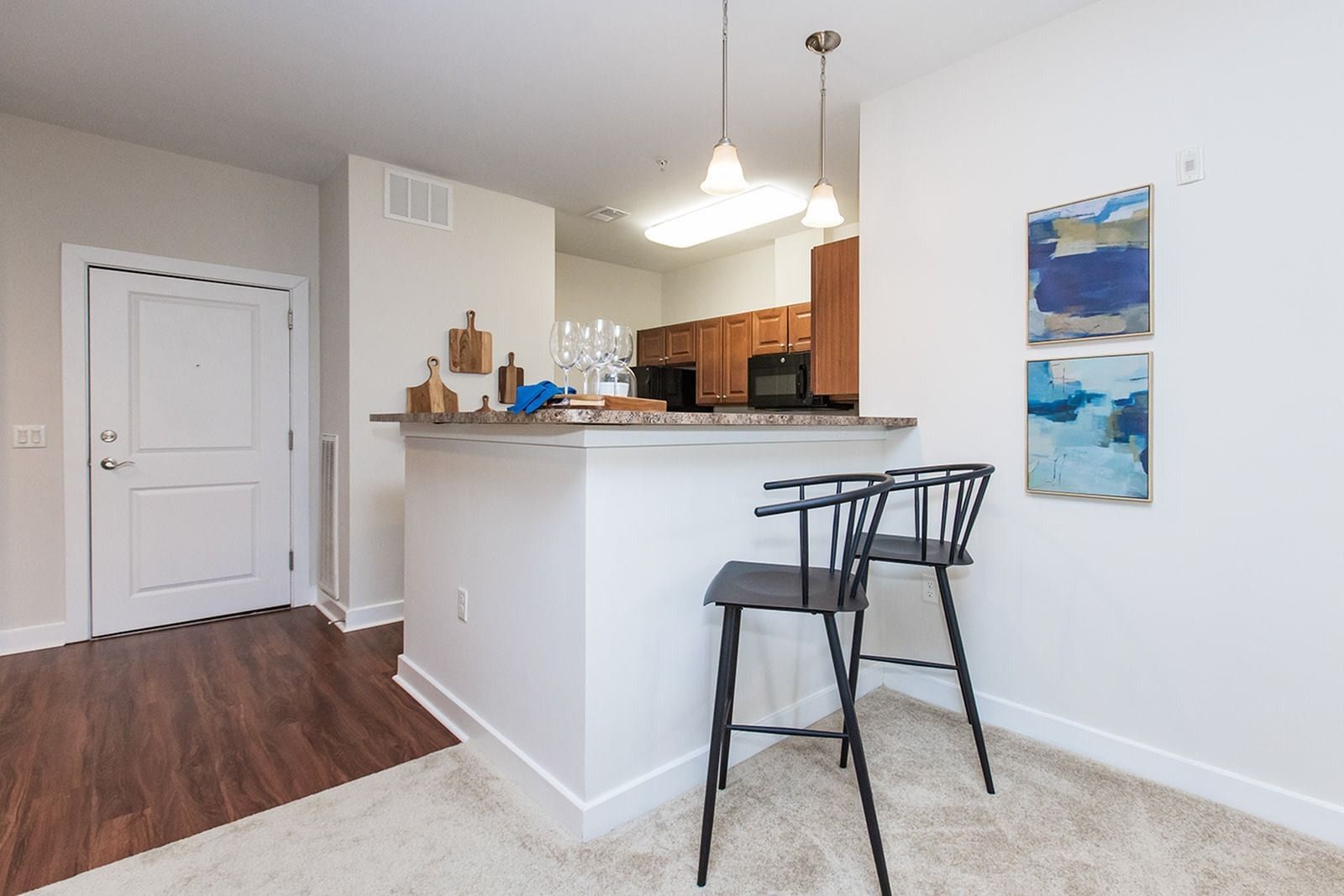 Open-concept kitchen and living area with white walls, bar stools, and hardwood and carpet flooring at Verde at Howard Square West Apartments in Elkridge, MD.