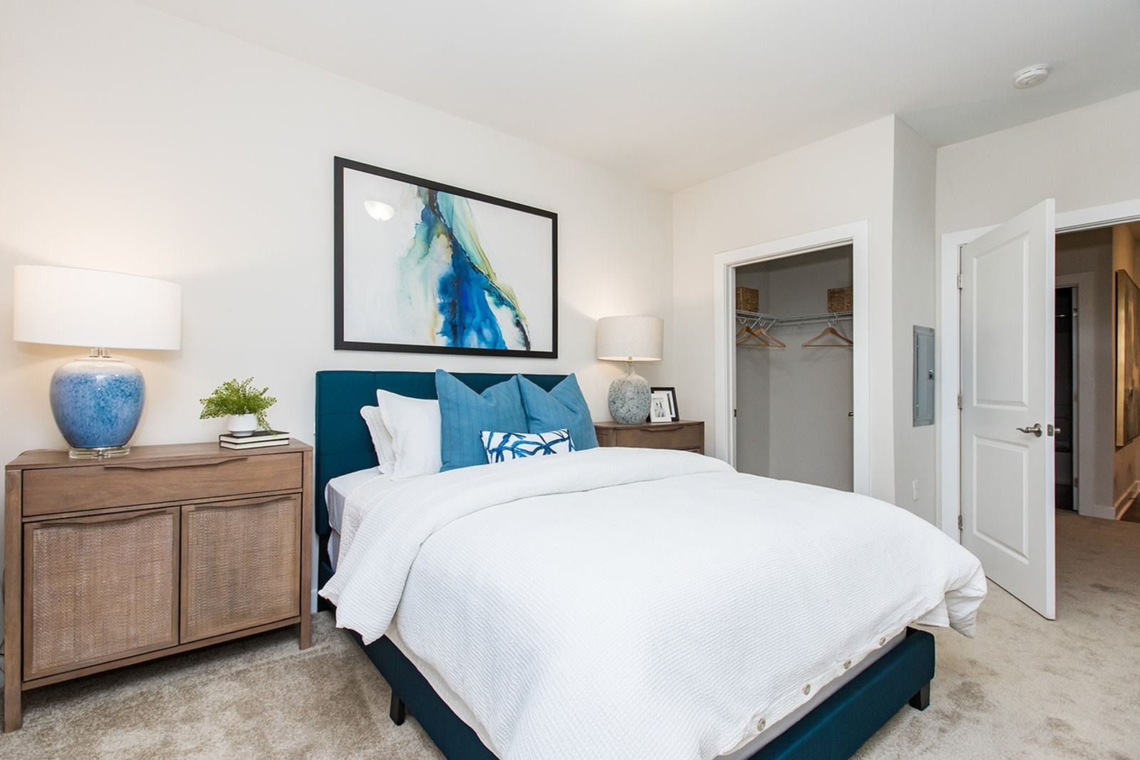 Bright bedroom with teal upholstered bed, white bedding, woven nightstand, and abstract wall art at Verde at Howard Square West Apartments in Elkridge, MD.