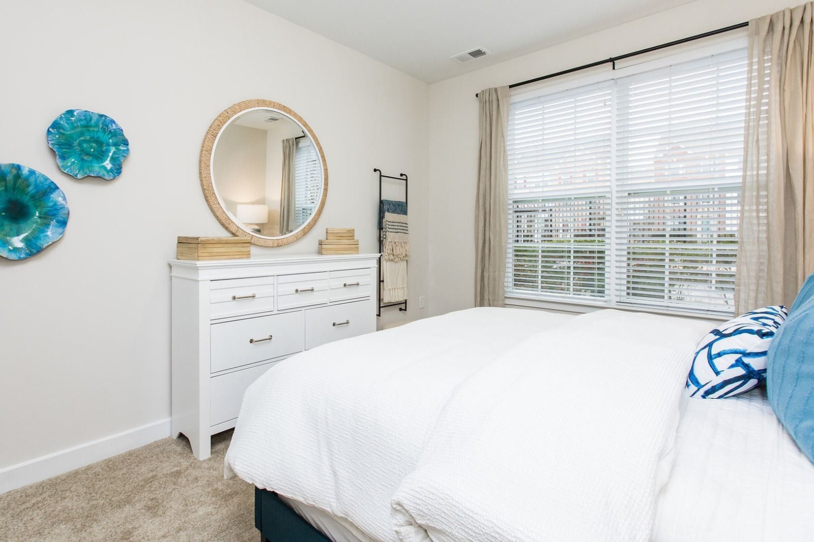 Bright bedroom with white bedding, vanity, round mirror, teal wall decor, and large window blinds at Verde at Howard Square West Apartments in Elkridge, MD.