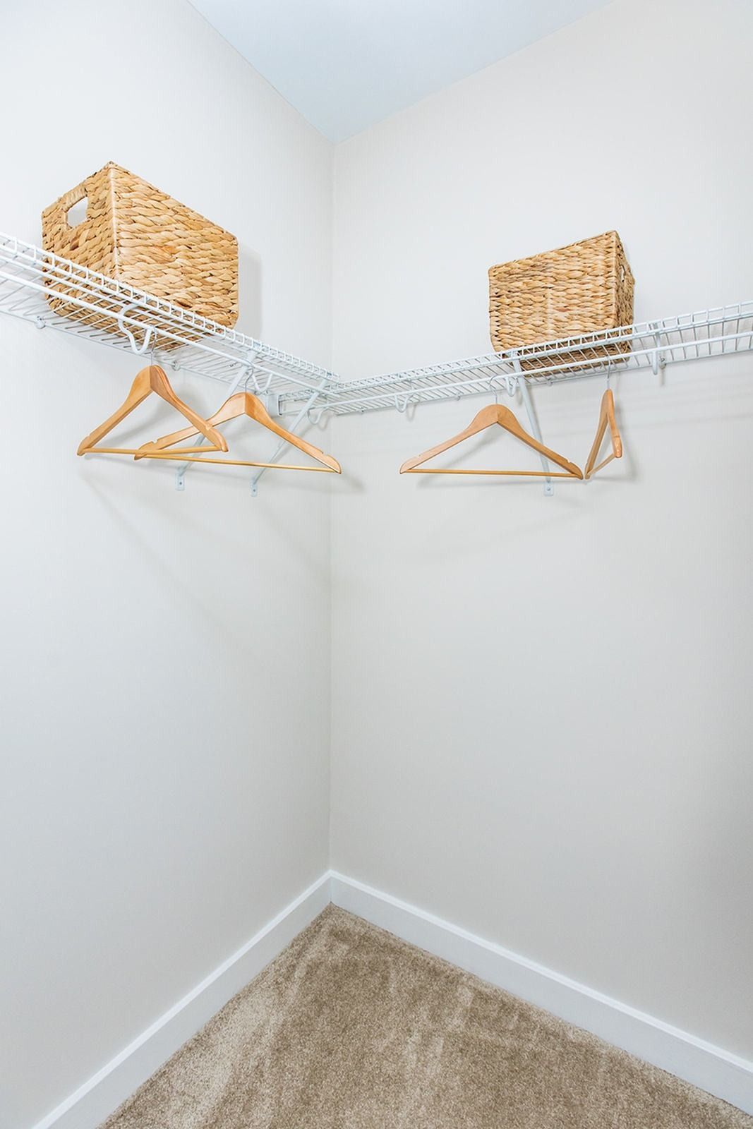 Empty closet corner with wire shelves, wooden hangers, woven baskets, and beige carpet at Verde at Howard Square West Apartments in Elkridge, MD.