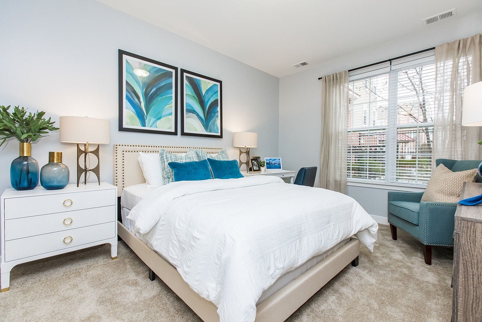 Bright modern bedroom with white bed, blue accents, nightstands, and a window seat by large windows at Verde at Howard Square West Apartments in Elkridge, MD.