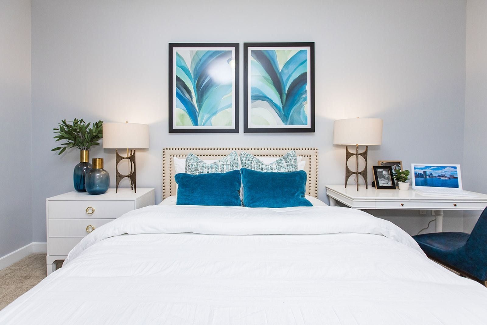 Bright bedroom with white bed, blue accents, framed teal artwork, and matching bedside lamps at Verde at Howard Square West Apartments in Elkridge, MD.