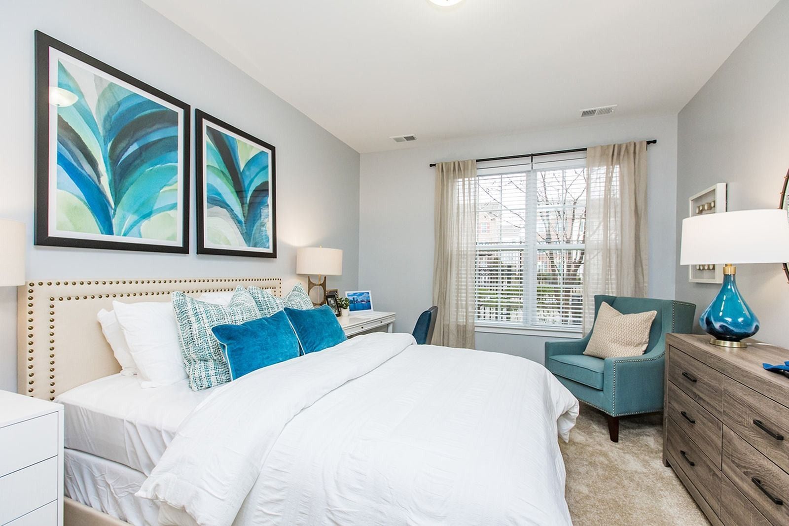 Bright modern bedroom with white bedding, teal accents, framed art, and a window seat at Verde at Howard Square West Apartments in Elkridge, MD.
