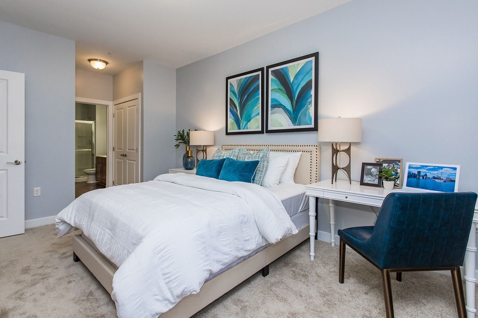 Bright bedroom with a white bed, blue accents, wall art, and a small desk by the window at Verde at Howard Square West Apartments in Elkridge, MD.