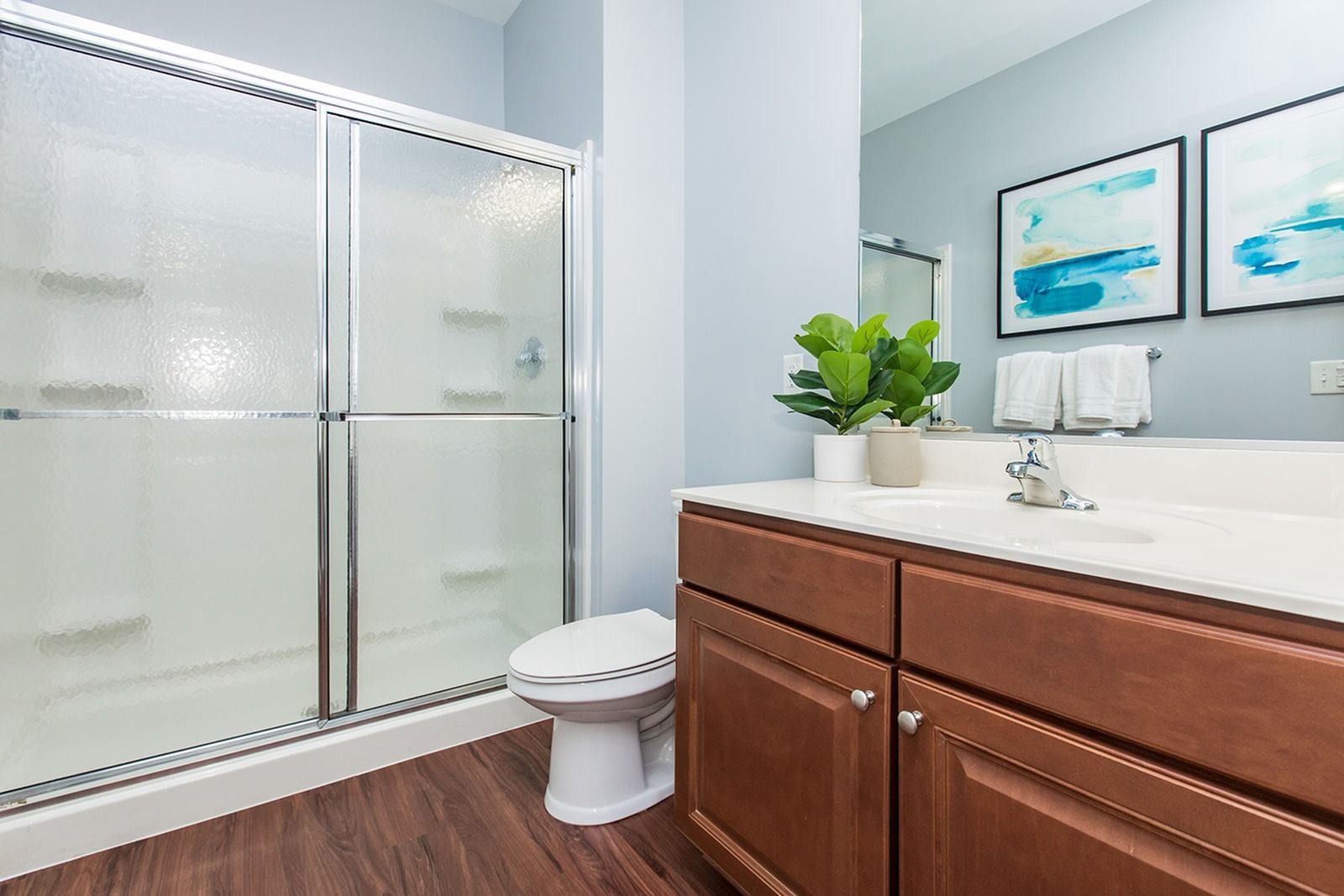 Modern bathroom with glass shower, wooden vanity, white fixtures, and blue wall art at Verde at Howard Square West Apartments in Elkridge, MD.