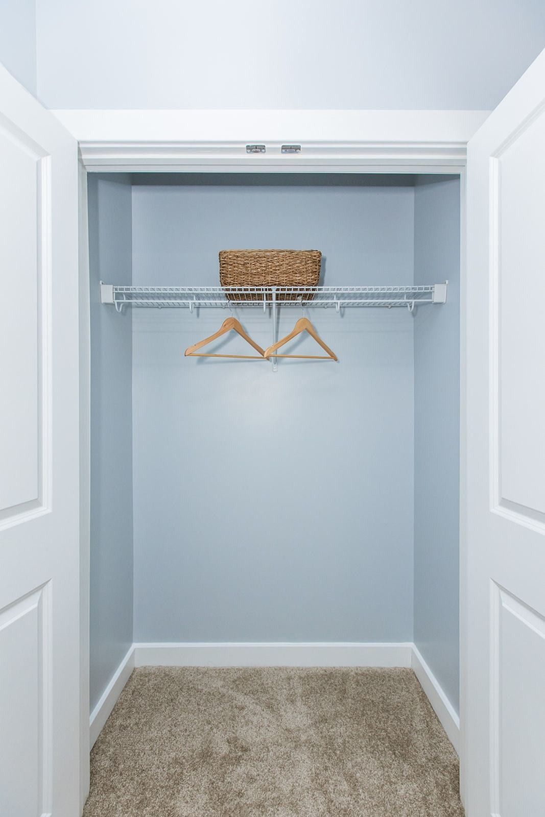 Empty closet with light blue walls, wire shelf, two hangers, and a woven basket on the top shelf at Verde at Howard Square West Apartments in Elkridge, MD.