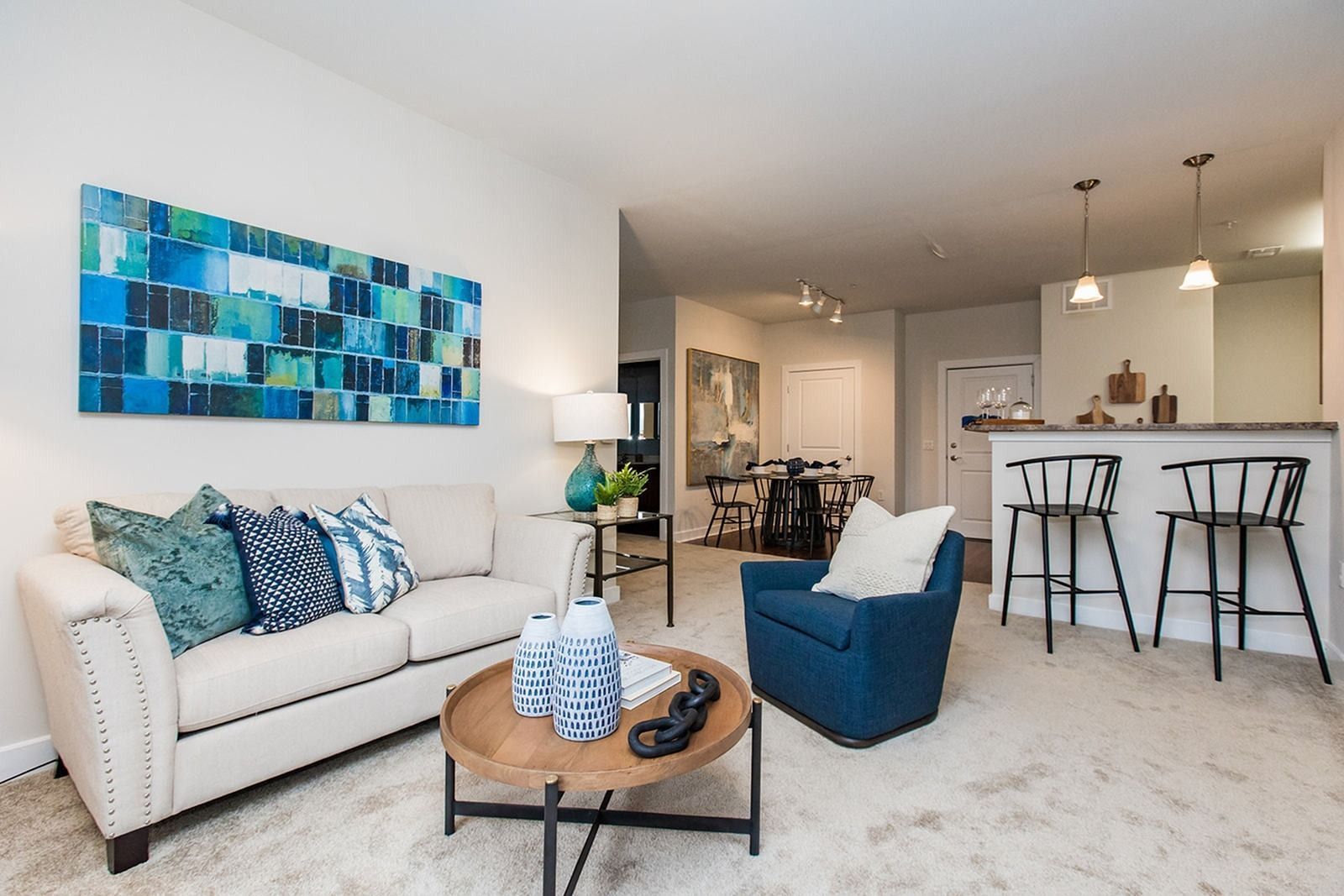 Bright modern living room with white sofa, blue accents, coffee table, and open kitchen with bar stools at Verde at Howard Square West Apartments in Elkridge, MD.