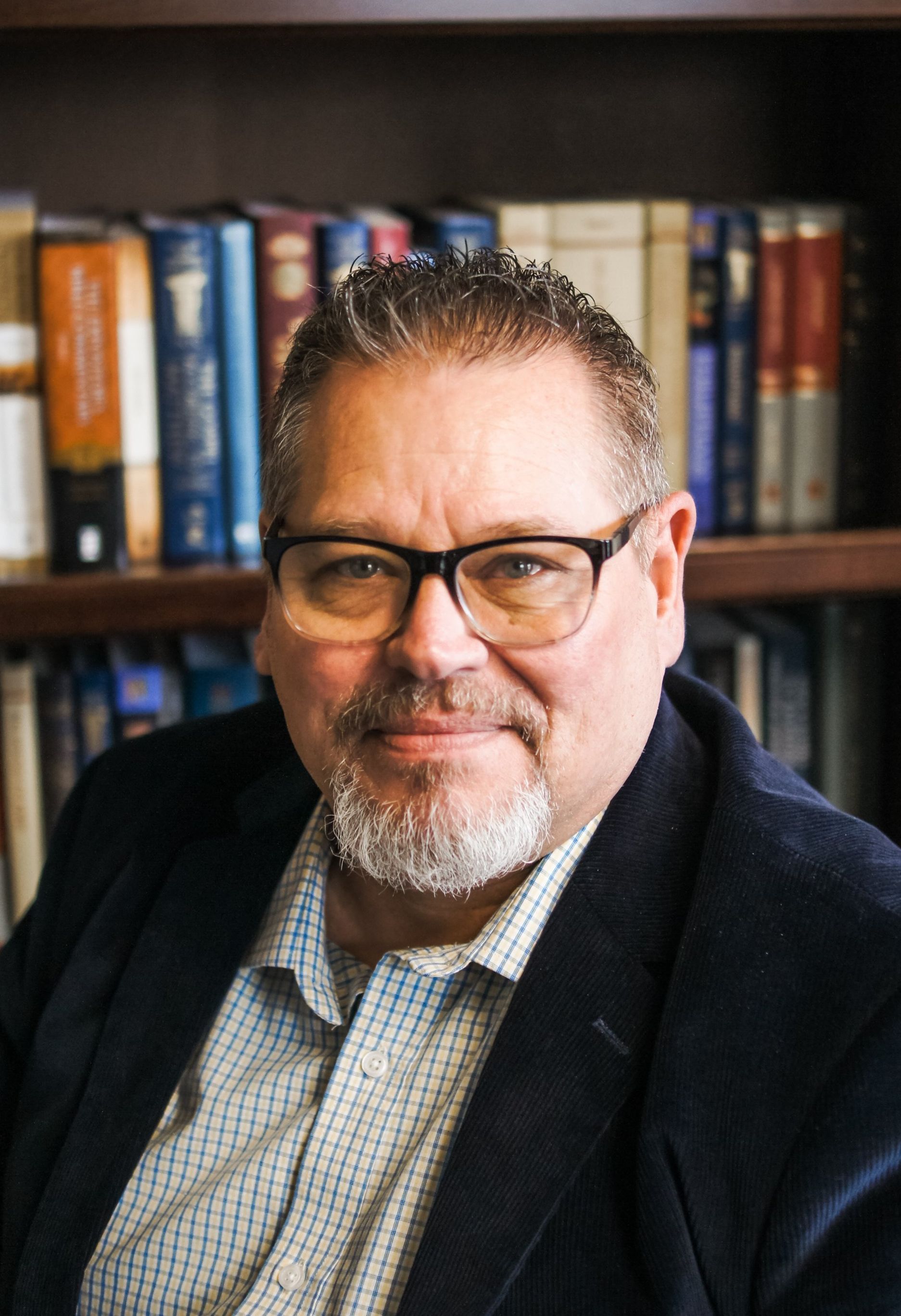 A man with glasses and a beard is sitting in front of a bookshelf.