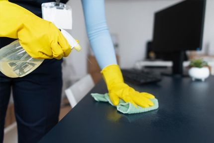 A person wearing yellow gloves is cleaning a desk with a cloth and spray bottle.