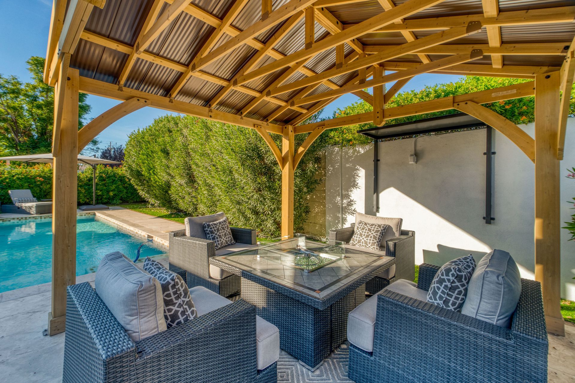 Outdoor seating area under a wooden gazebo with pool view. Wicker furniture, glass-topped table, and lush greenery.