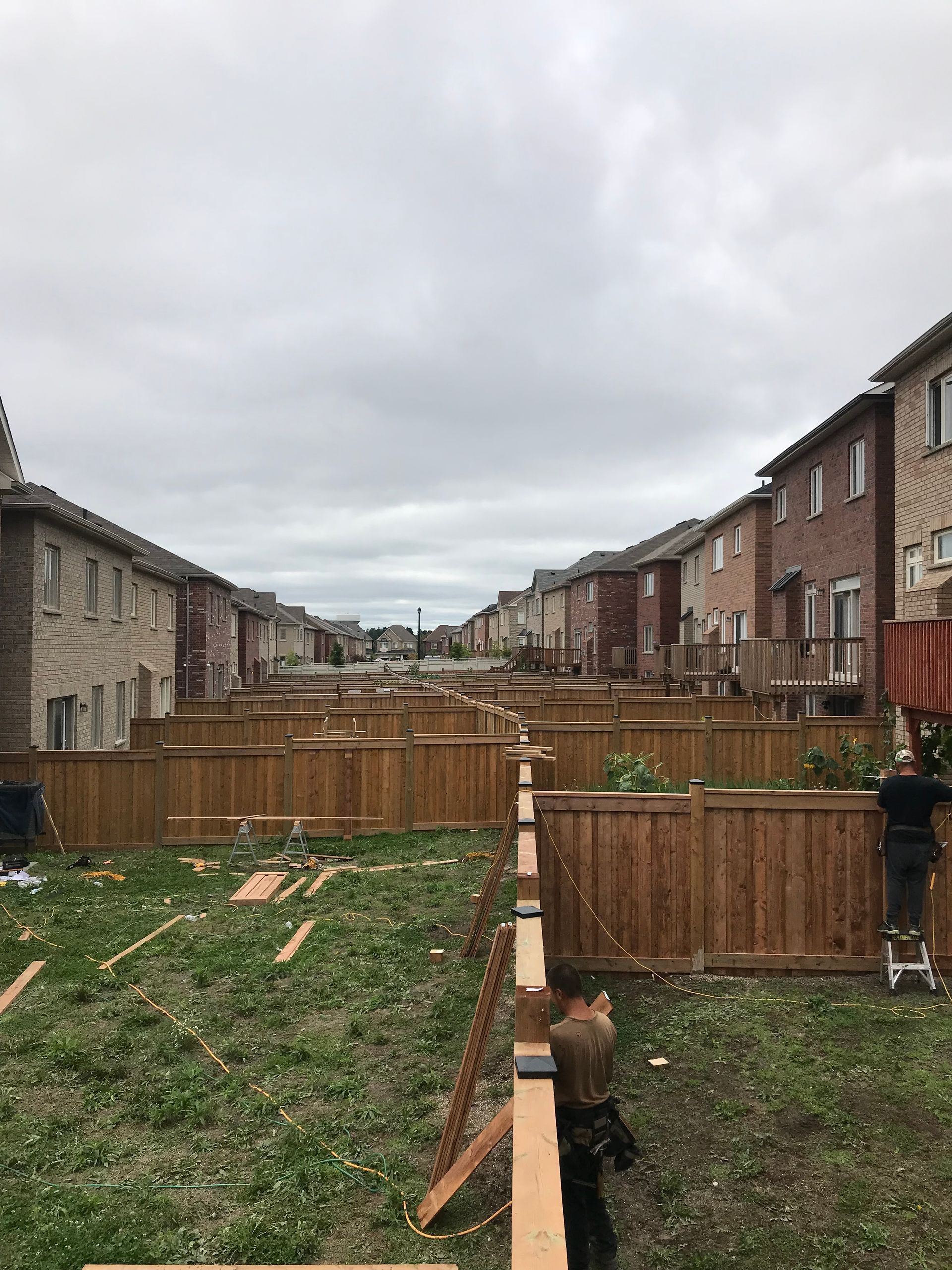 Workers building a wooden fence in a suburban backyard, with rows of houses on either side, cloudy sky.