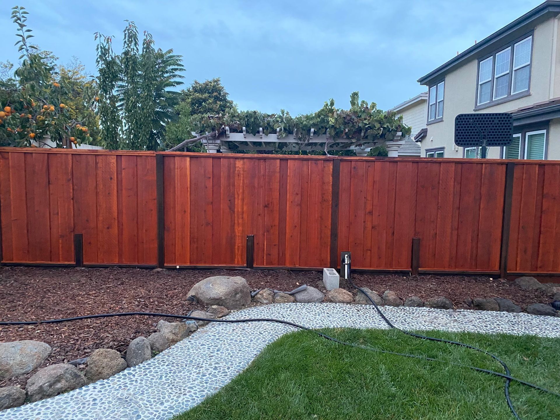 A wooden fence in a backyard with a grass lawn and a stone walkway.