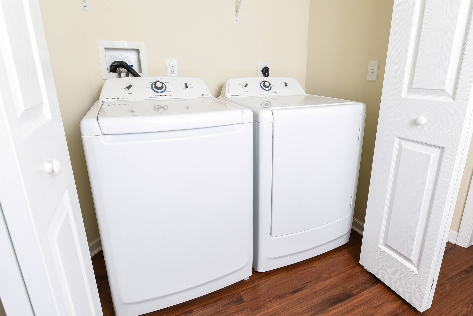 White washer and dryer side by side in a small laundry closet with wood floor.