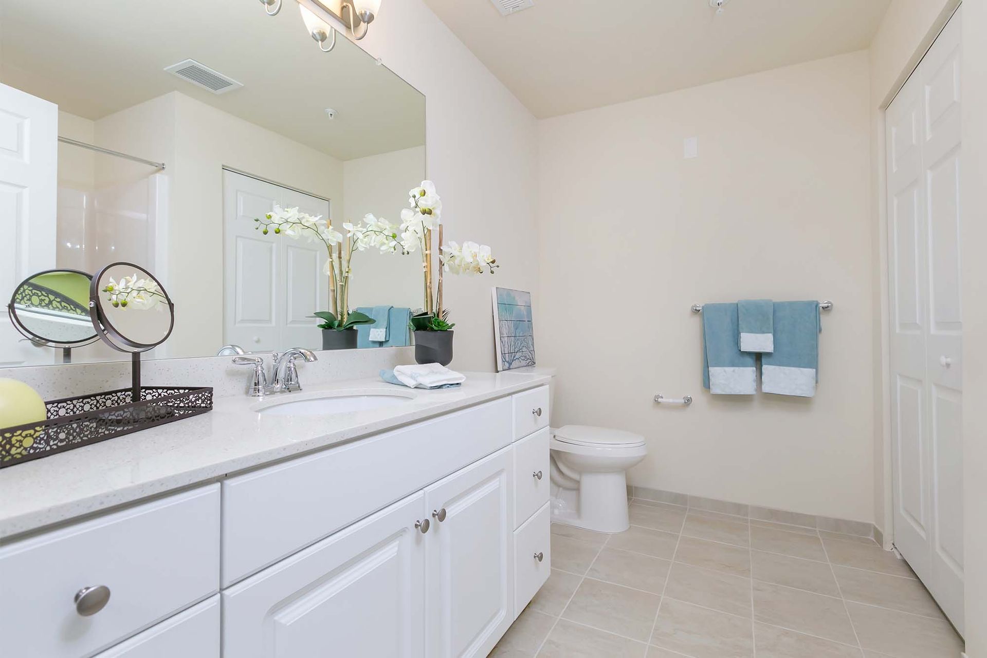 Bright bathroom in an apartment featuring a long white vanity, large mirror, toilet, and teal towels.