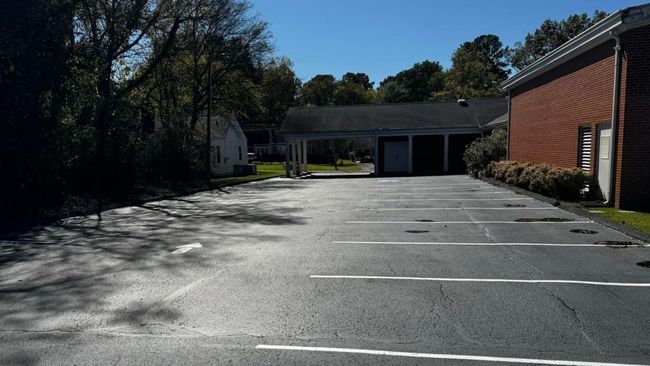 Empty asphalt parking lot with white lines, trees, and buildings on a sunny day.