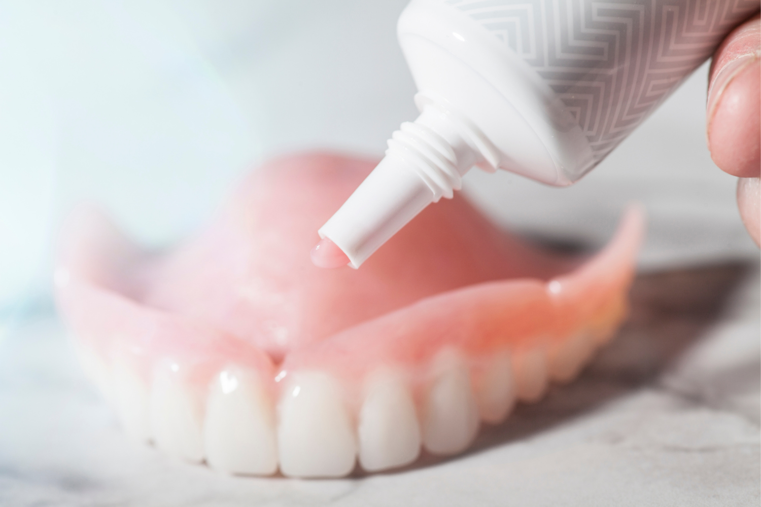 A person applying adhesive cream from a tube onto the surface of a set of pink dentures.