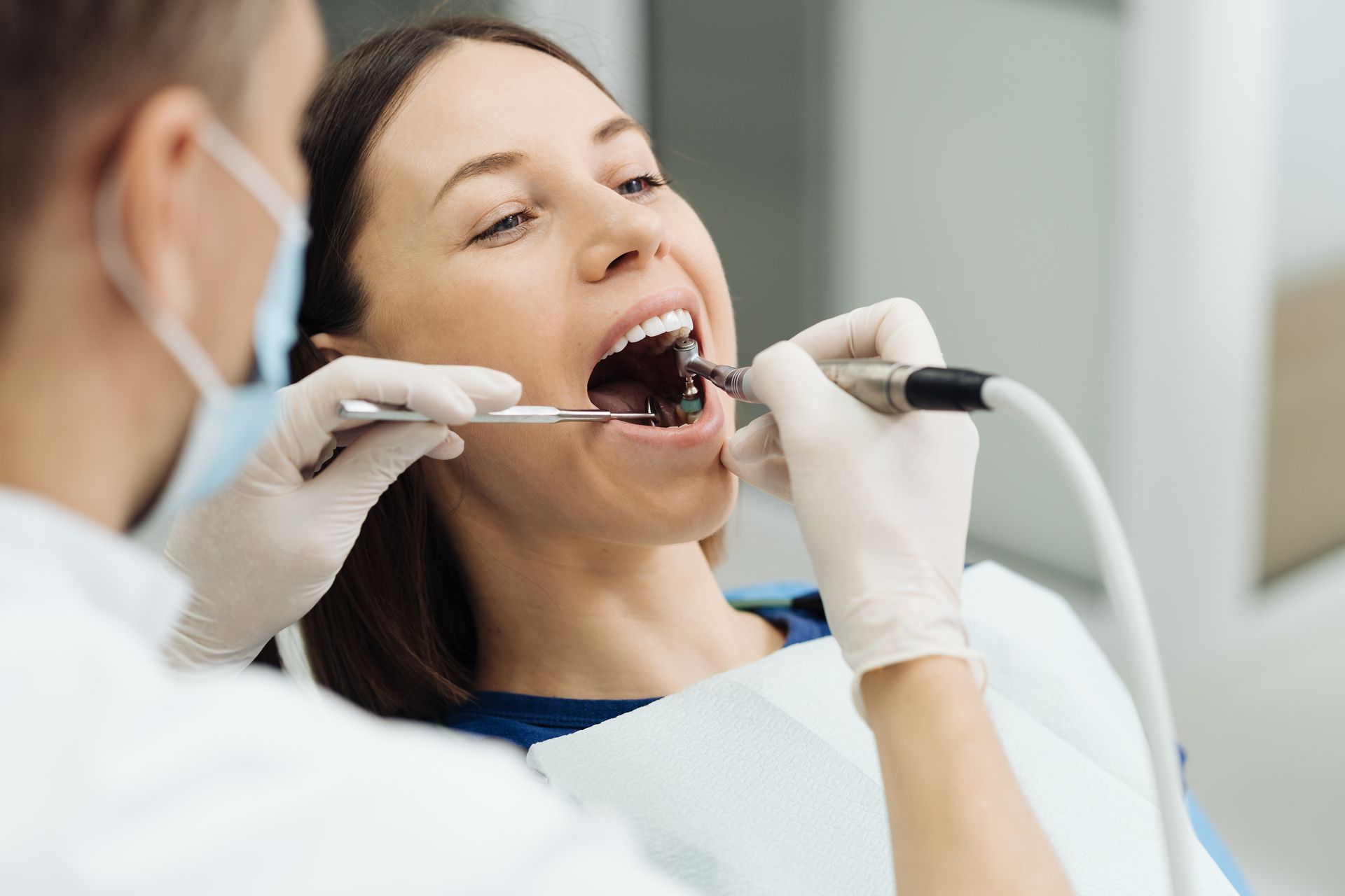 A dental professional in a mask and gloves uses a dental handpiece to perform a procedure on a patient's open mouth.