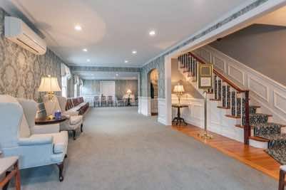 An elegant, carpeted funeral home waiting area with chairs, wallpaper, and a staircase.