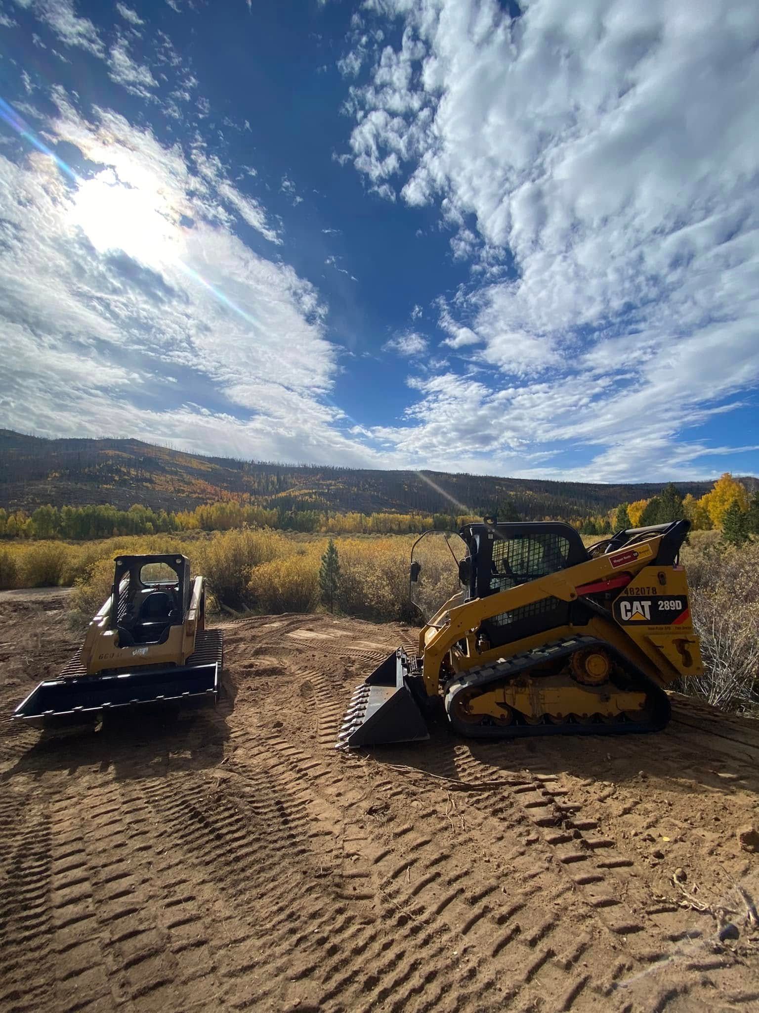 Two excavators on a dirt lot under a cloudy blue sky, with hills in the background.