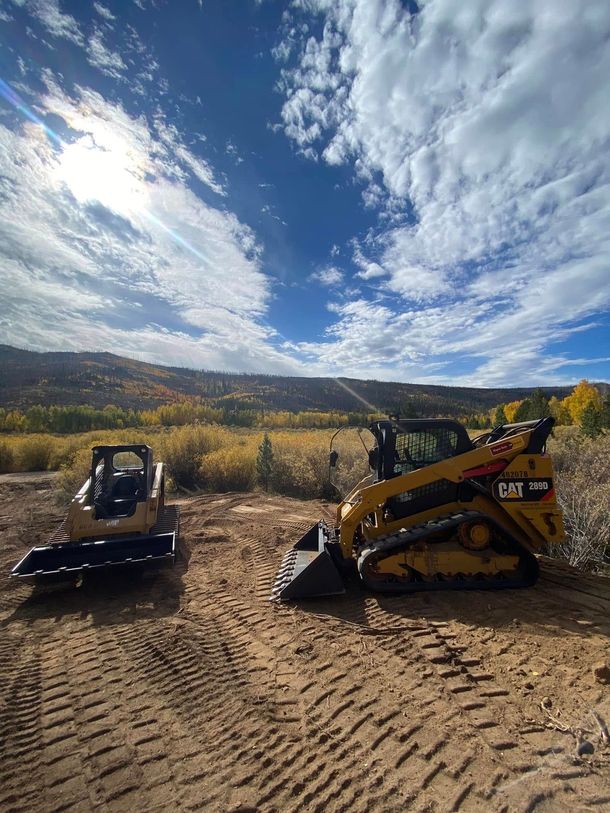 Two small excavators on a dirt lot under a bright, cloudy sky with mountains in the background