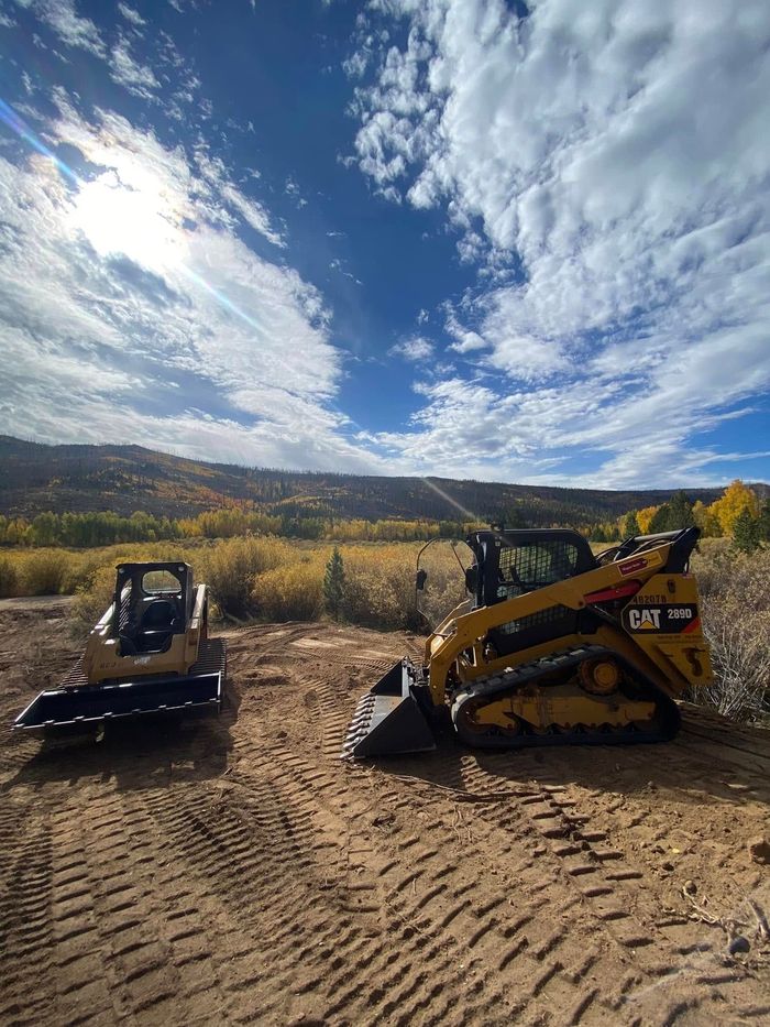 Two small excavators on a dirt lot under a bright, cloudy sky with mountains in the background