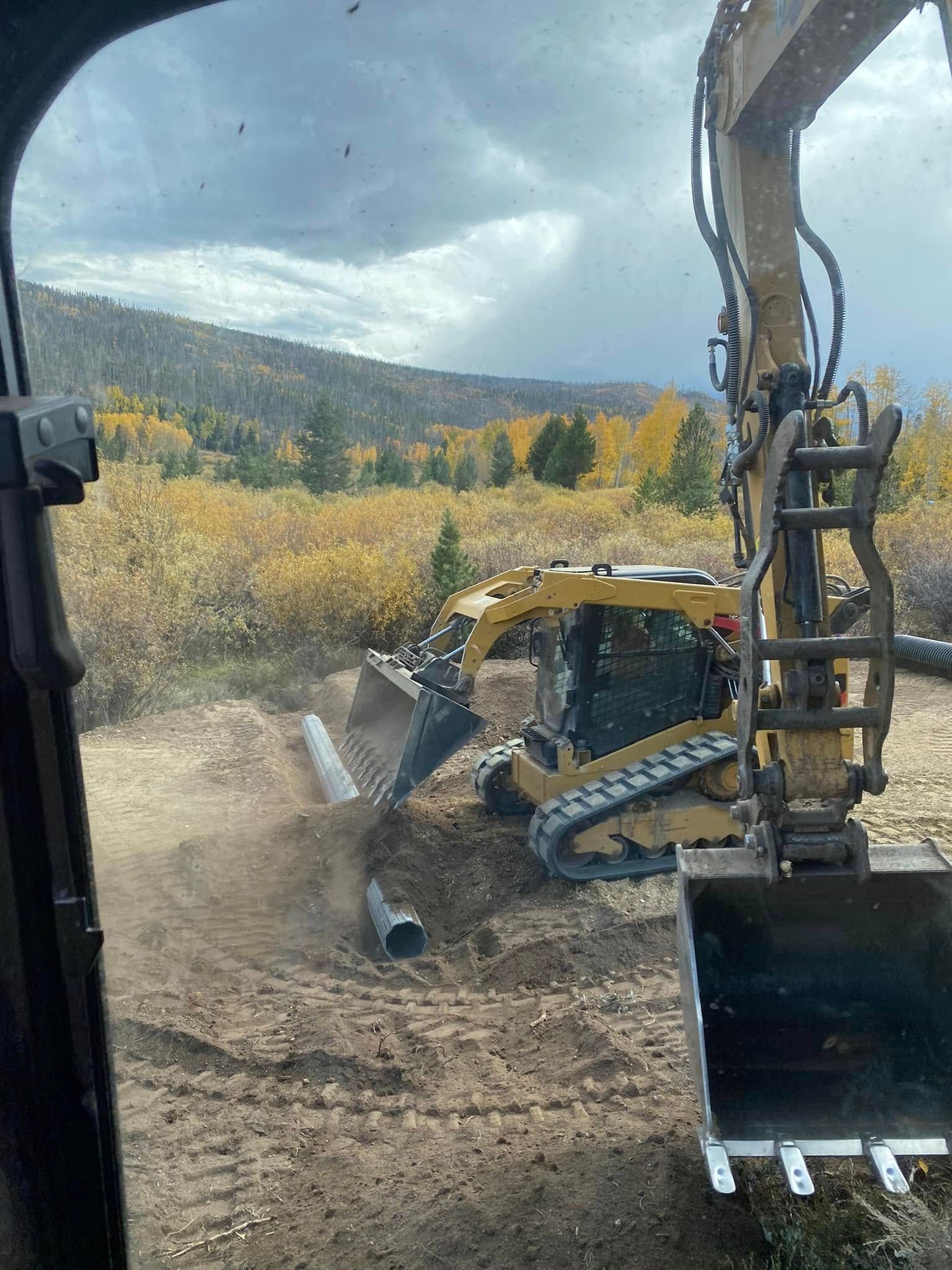 Excavator clearing dirt beside a forested hillside under cloudy skies, with a dust cloud rising.