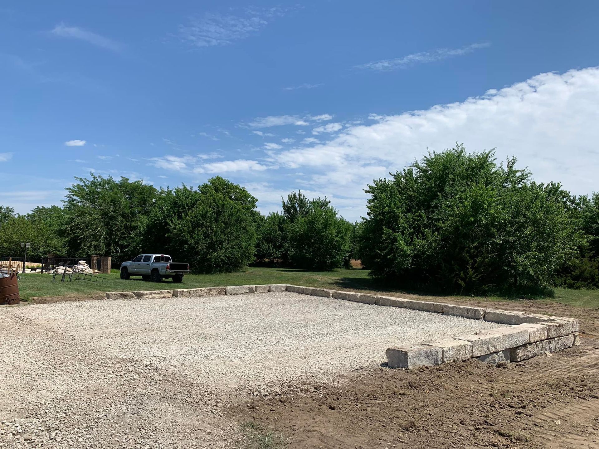 Gravel lot beside a stone curb with trees and a parked pickup truck under a partly cloudy sky