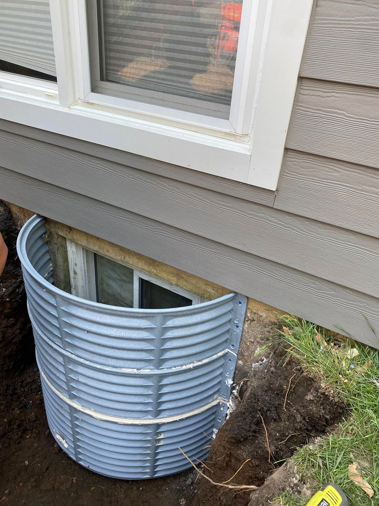 Corrugated metal window well beside a house foundation with a basement window and exposed soil.