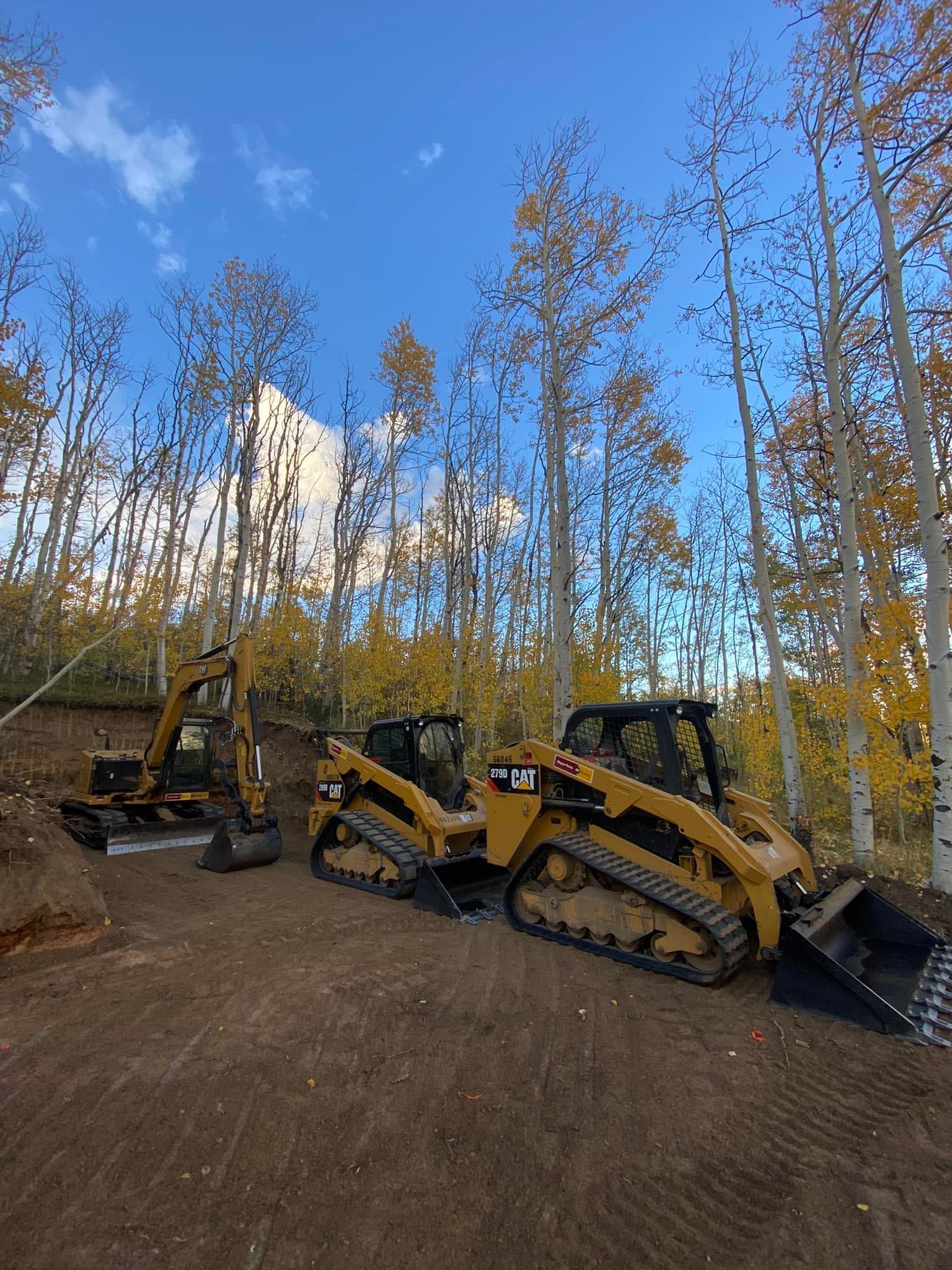 Two yellow tracked construction machines parked on a dirt lot in a leafless forest under a blue sky.