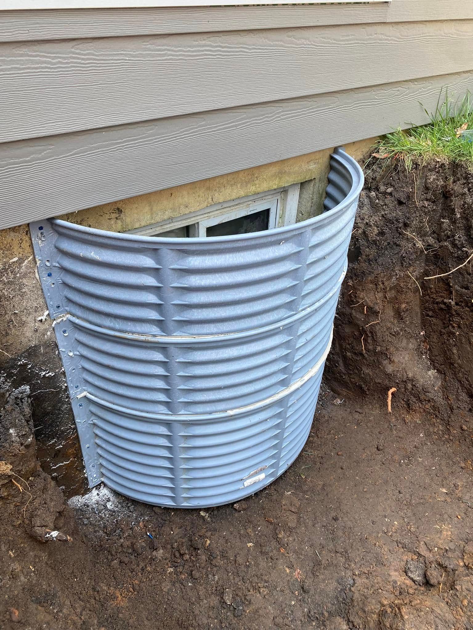Blue corrugated window well covering a basement window beside a house foundation and dirt trench