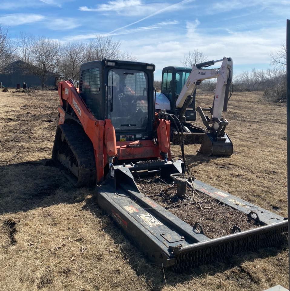 Orange skid steer with a brush mower attachment beside a small excavator in a dry field.