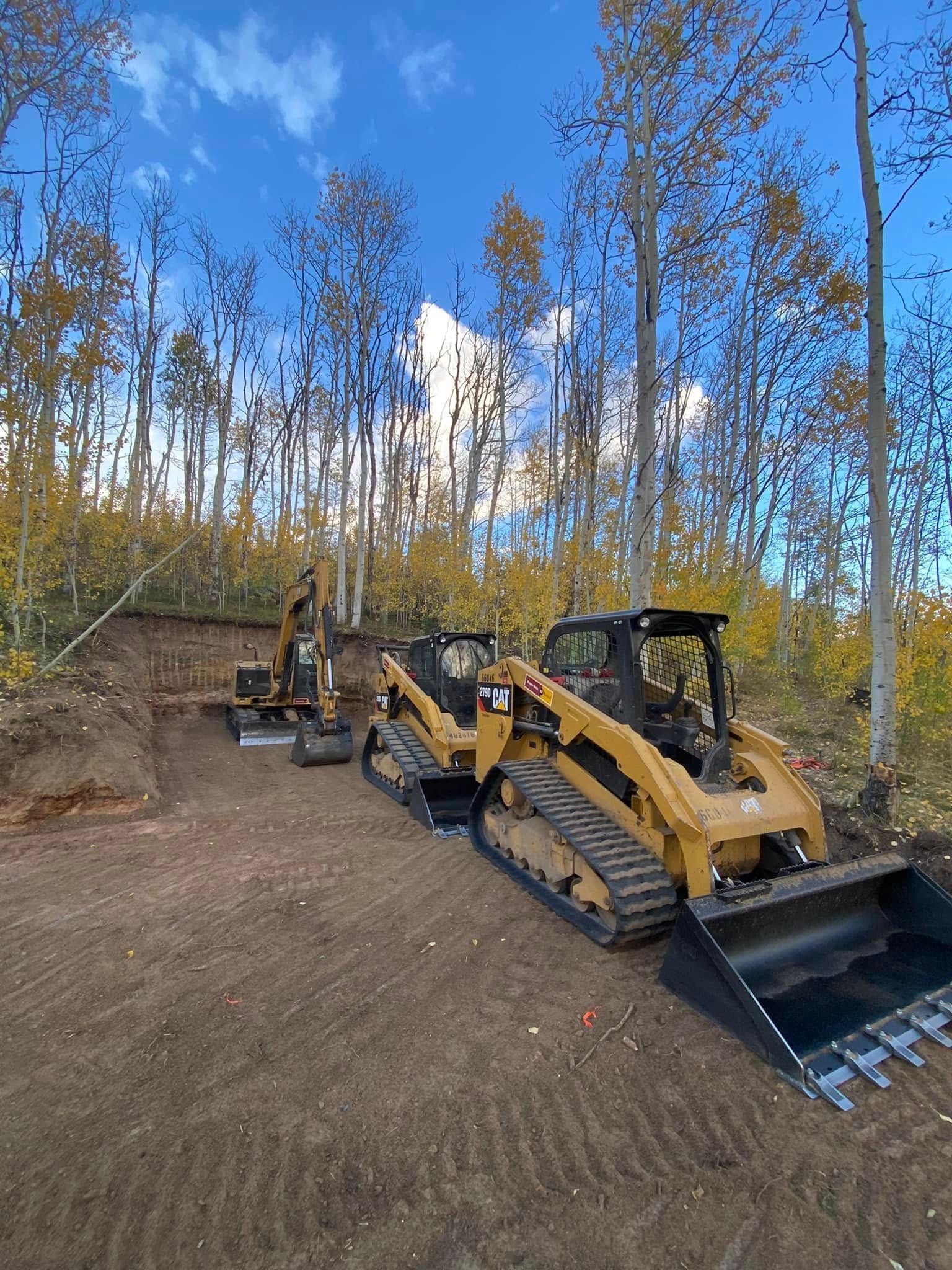 Yellow tracked skid steer and excavator on a dirt slope in a forest clearing under blue sky